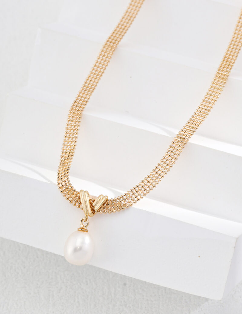 A gold necklace (product: o_1i0fcngqa46hech98l1l221c7f1) with a delicate chain and a single, large, teardrop-shaped pearl pendant is displayed on a white, textured background. The chain features intricate lace-like links in 18k Gold Vermeil, adding to its elegance, and the pearl has a smooth, lustrous surface. Elegant pearl jewelry