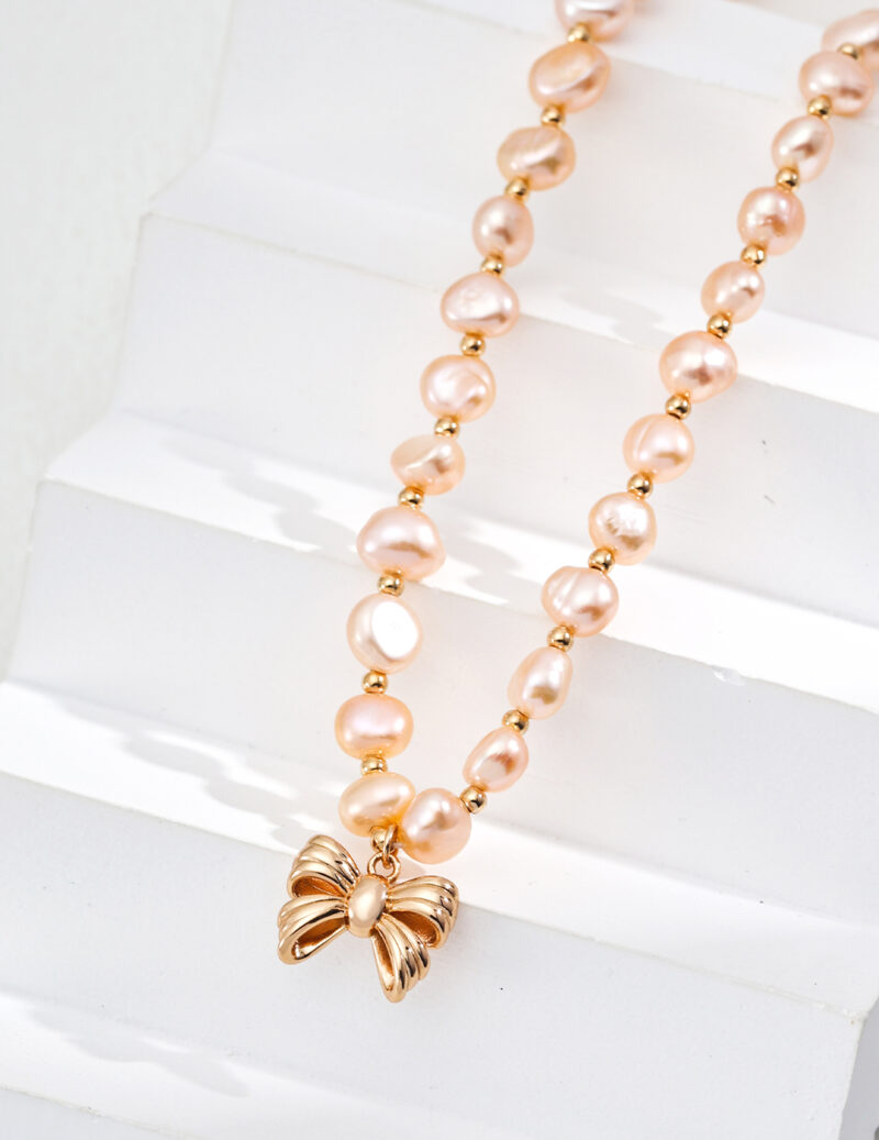 The product, identified as o_1ia1s7qhq18cu26mcbmgsmuht1, is a stunning pearl necklace featuring irregularly shaped pink pearls combined with small 18k gold vermeil beads. This elegant piece is showcased against a white step background and is beautifully finished with a graceful gold bowtie-shaped pendant at its center. Elegant pearl jewelry