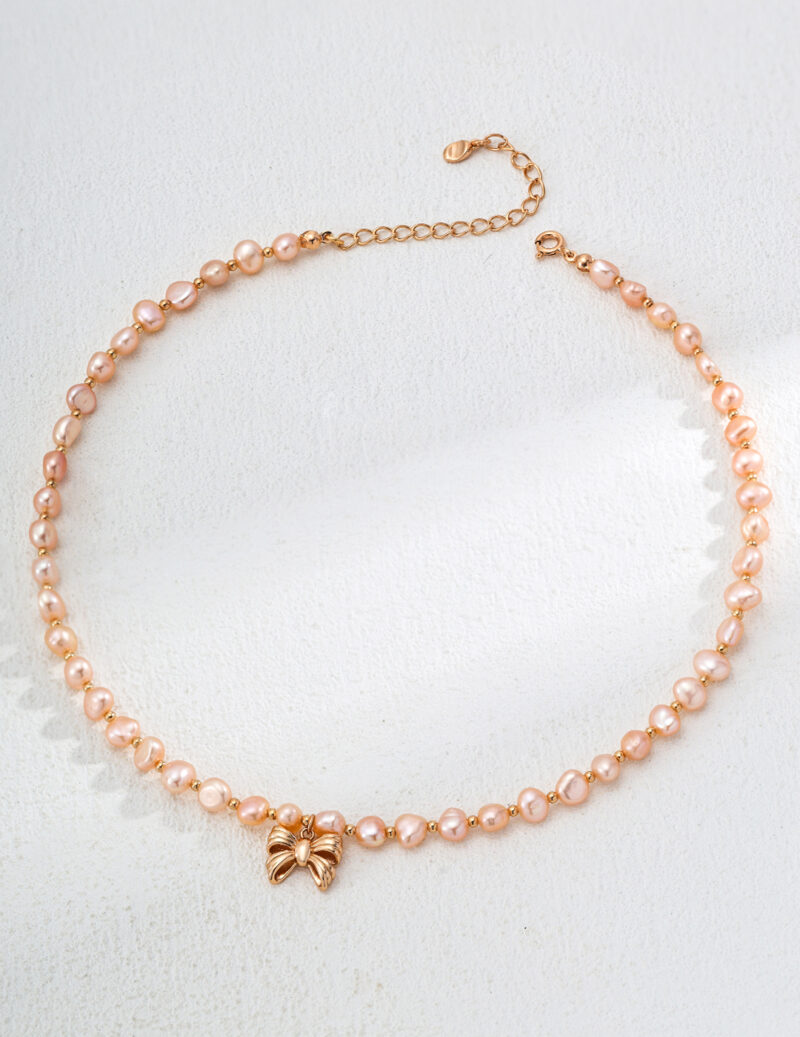 The o_1ia1s7qhr11ld1e0ett91jgemk52, a delicate pearl necklace with small pink pearls beautifully linked together, is accented by a gold vermeil butterfly charm. Displayed in a circular arrangement on a light, textured background, this elegant piece embodies the grace of a perfectly tied bowtie. Elegant pearl jewelry