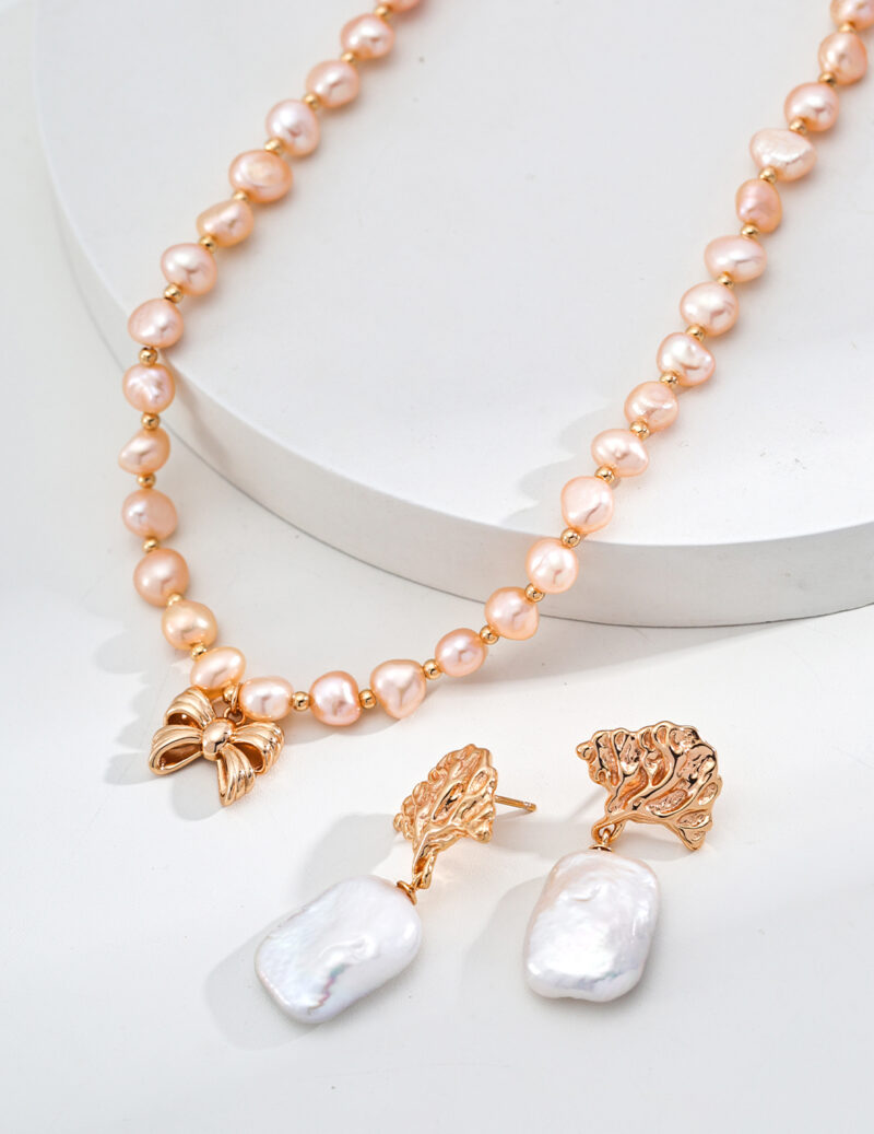 The product o_1ia1s7qhr9do1g2912hl100411f33 features a pearl necklace with an 18k gold vermeil bow, elegantly displayed on a white surface. Below the necklace, a pair of earrings highlight gold, leaf-inspired settings, each embellished with large, irregular white pearls. Elegant pearl jewelry