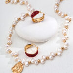 The o_1ia52bhcic7pdtpbi1cm41461b features a beaded necklace with alternating white pearls and gold beads, adorned with a gold pendant, artfully displayed on a smooth white stone. Accompanying the display is a pair of gold hoop earrings accented with red tones. Elegant pearl jewelry