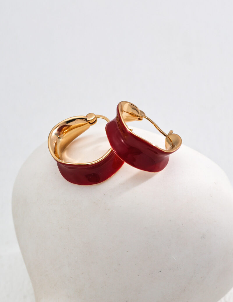 The product o_1ia52bhcj8uo10ge1toq1t1v1al5d consists of a pair of red enamel earrings, elegantly presented on a white, rounded surface. The gold hoops have a smooth, shiny finish that creates a beautiful contrast against the matte background. Elegant pearl jewelry