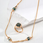 Product o_1ia9hugl1emtpnrk1s12pj1qc22, a gold necklace adorned with green stone beads accompanied by a matching gold ring featuring a green stone, is elegantly presented on a textured white surface. Elegant pearl jewelry