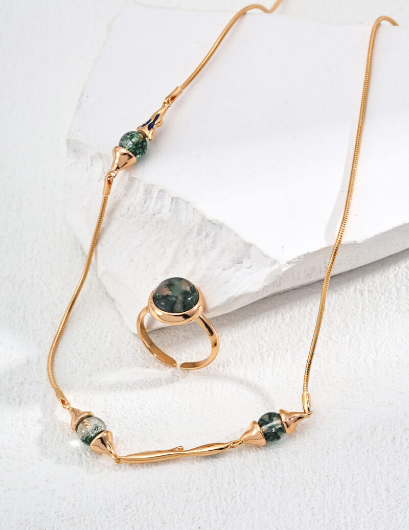 Product o_1ia9hugl1emtpnrk1s12pj1qc22, a gold necklace adorned with green stone beads accompanied by a matching gold ring featuring a green stone, is elegantly presented on a textured white surface. Elegant pearl jewelry