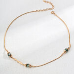 The o_1ia9hugl21vim1d46g9pkcui445 product is an elegant gold necklace featuring three small, uniquely shaped green stones arranged along the chain. It is showcased against a gently textured light background. Elegant pearl jewelry