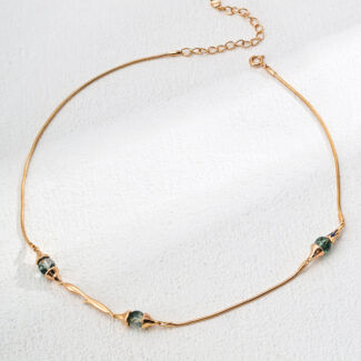 The o_1ia9hugl21vim1d46g9pkcui445 product is an elegant gold necklace featuring three small, uniquely shaped green stones arranged along the chain. It is showcased against a gently textured light background. Elegant pearl jewelry