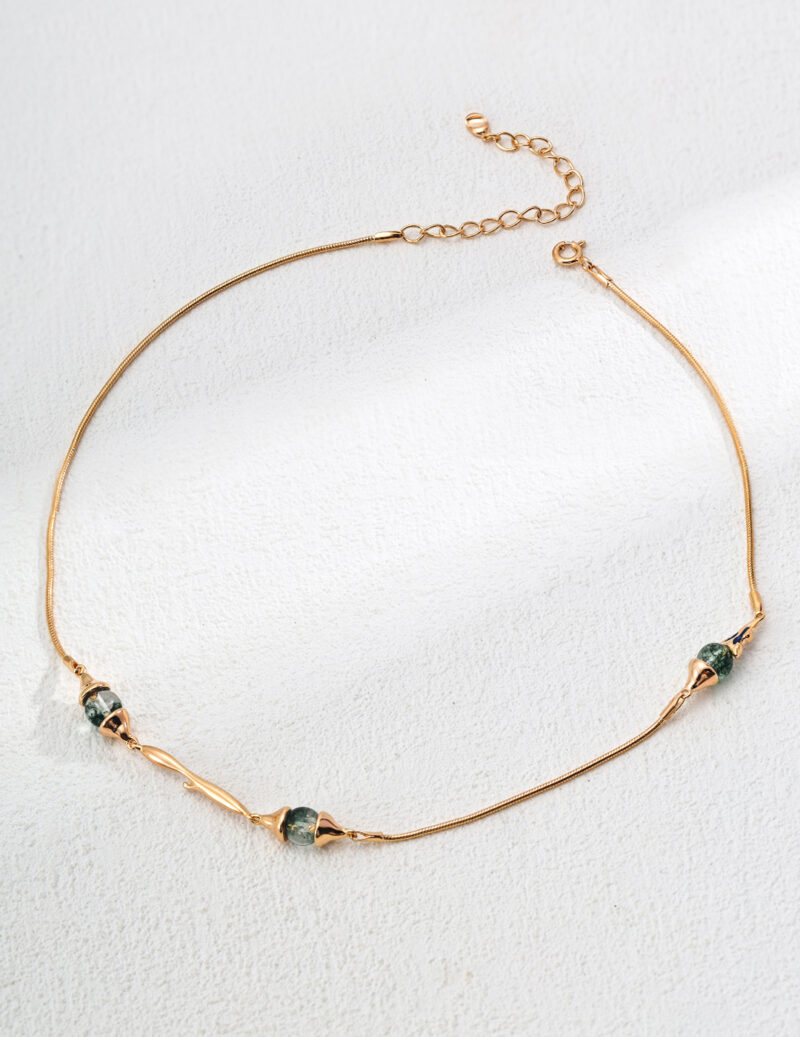 The o_1ia9hugl21vim1d46g9pkcui445 product is an elegant gold necklace featuring three small, uniquely shaped green stones arranged along the chain. It is showcased against a gently textured light background. Elegant pearl jewelry