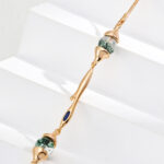 A delicate gold bracelet adorned with two green marbled beads reminiscent of Moss Agate and a small blue enamel accent, beautifully presented on white stepped surfaces. Elegant pearl jewelry