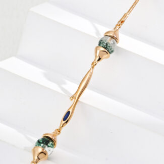 A delicate gold bracelet adorned with two green marbled beads reminiscent of Moss Agate and a small blue enamel accent, beautifully presented on white stepped surfaces. Elegant pearl jewelry