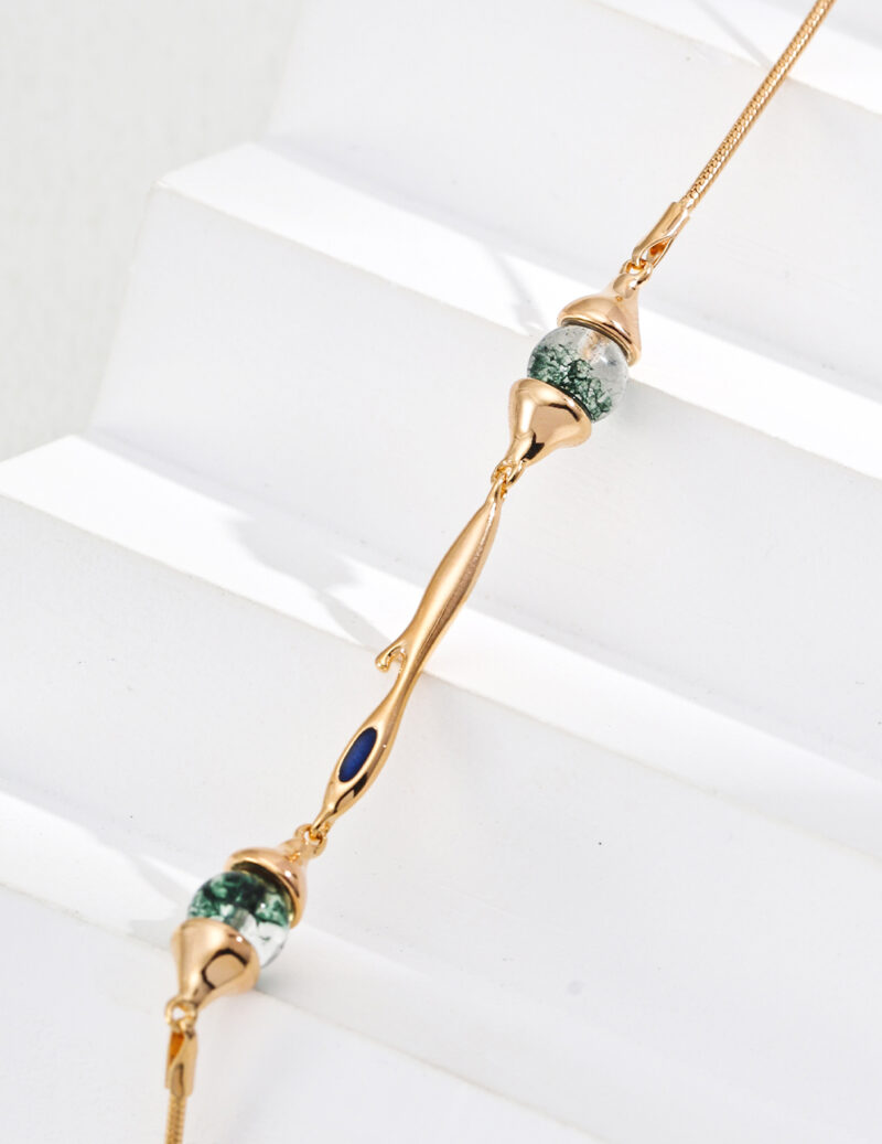 A delicate gold bracelet adorned with two green marbled beads reminiscent of Moss Agate and a small blue enamel accent, beautifully presented on white stepped surfaces. Elegant pearl jewelry