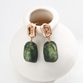 Elegant minimalist gold-toned earrings featuring large, glossy green stone pendants from the o_1iac7l2s6fkp1fq2j63tqsmsq1 collection rest gracefully on a small, white ceramic vase. These abstract earrings skillfully merge metallic and natural elements against a textured white background, evoking the charm of faux amber. Elegant pearl jewelry