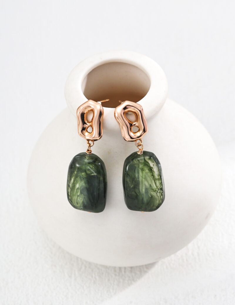 Elegant minimalist gold-toned earrings featuring large, glossy green stone pendants from the o_1iac7l2s6fkp1fq2j63tqsmsq1 collection rest gracefully on a small, white ceramic vase. These abstract earrings skillfully merge metallic and natural elements against a textured white background, evoking the charm of faux amber. Elegant pearl jewelry