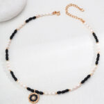 The o_1iac7vg141grn19jog88ha1r2p3 is displayed on a white surface next to a round, smooth stone. This necklace showcases alternating black onyx and white beads, accentuated by a gold pendant with an intricate design. Elegant pearl jewelry