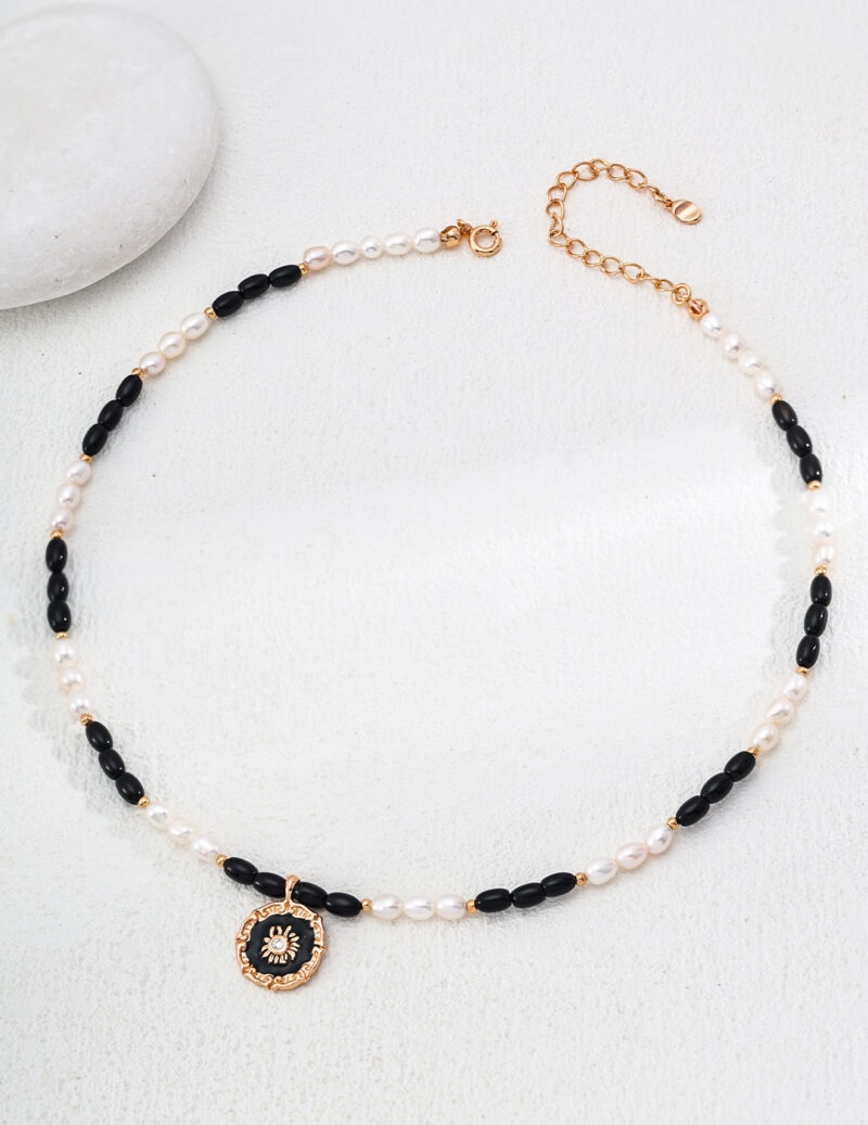 The o_1iac7vg141grn19jog88ha1r2p3 is displayed on a white surface next to a round, smooth stone. This necklace showcases alternating black onyx and white beads, accentuated by a gold pendant with an intricate design. Elegant pearl jewelry